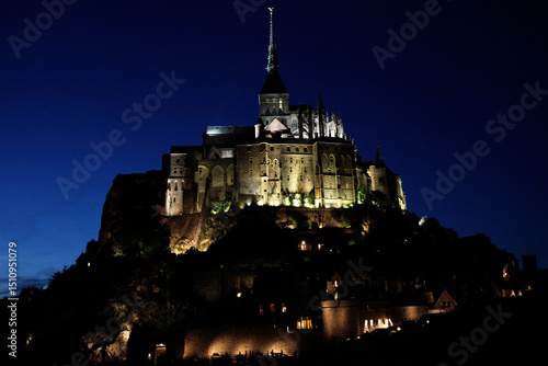 Fototapeta Naklejka Na Ścianę i Meble -  Night scene of Mont St Michel in France, with buildings lit by street lights and the tower lit by a search light, Normandy, northern France