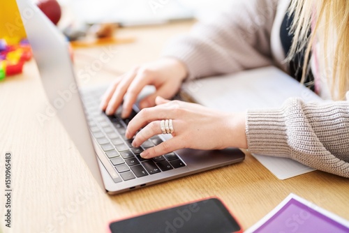 A woman is working on her laptop at a wooden desk. She's likely working from home, given the casual sweater and home setting.