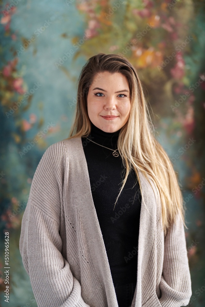 A young woman is posing for a portrait. She's wearing a black turtleneck and a beige cardigan, smiling softly against a floral backdrop. A young woman is posing for a portrait. She's wearing a black turtleneck and a beige cardigan, smiling softly against a floral backdrop.