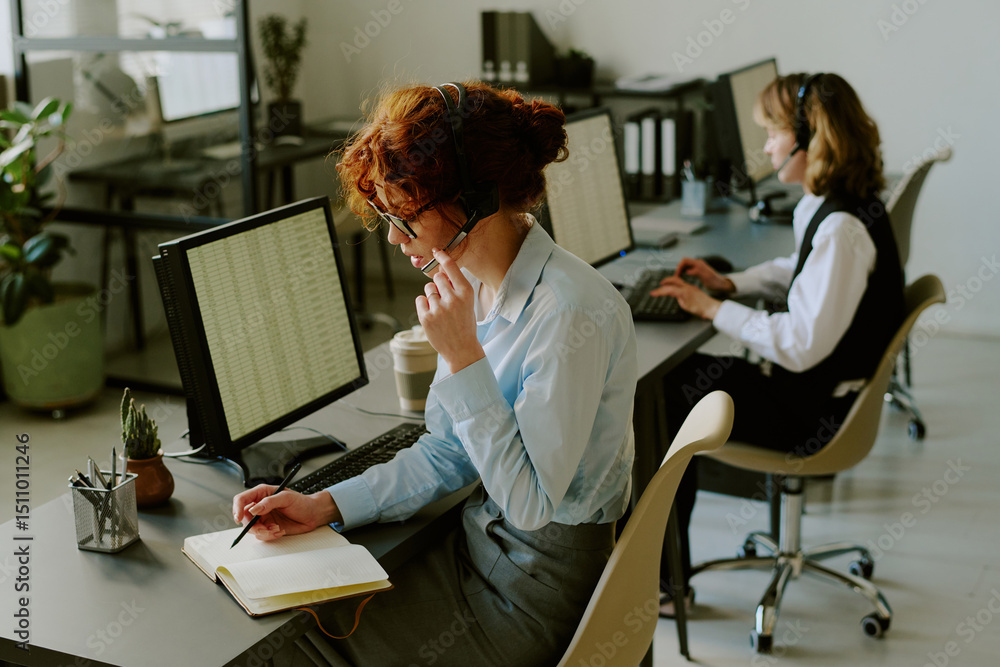 Obraz premium Two office workers focused on tasks at their desks, with one taking notes and wearing headset. Modern workspace with computers and plants creating productive environment