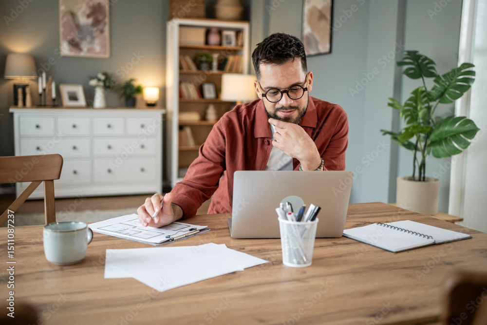 Focused architect working on blueprint at home office using laptop Focused architect working on blueprint at home office using laptop