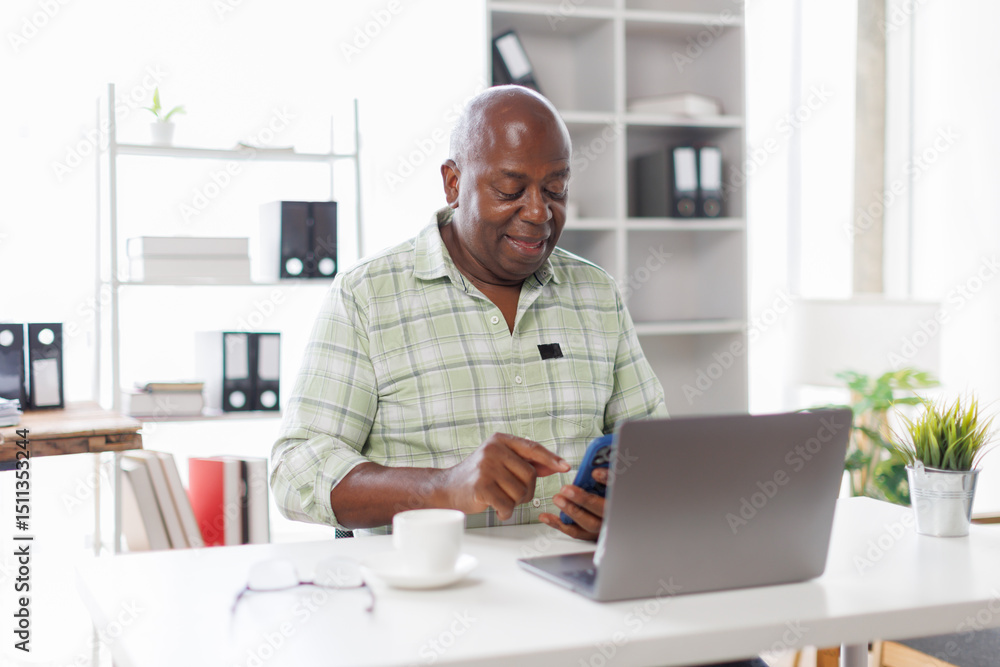 Senior businessman sitting alone in his home office and working on laptop computer. Smiling black man using laptop in living room. Businessman using phone laptop at home.
Senior businessman sitting alone in his home office and working on laptop computer. Smiling black man using laptop in living room. Businessman using phone laptop at home.
