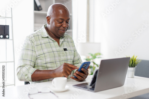 Wallpaper Mural Senior businessman sitting alone in his home office and working on laptop computer. Smiling black man using laptop in living room. Businessman using phone laptop at home.
 Torontodigital.ca