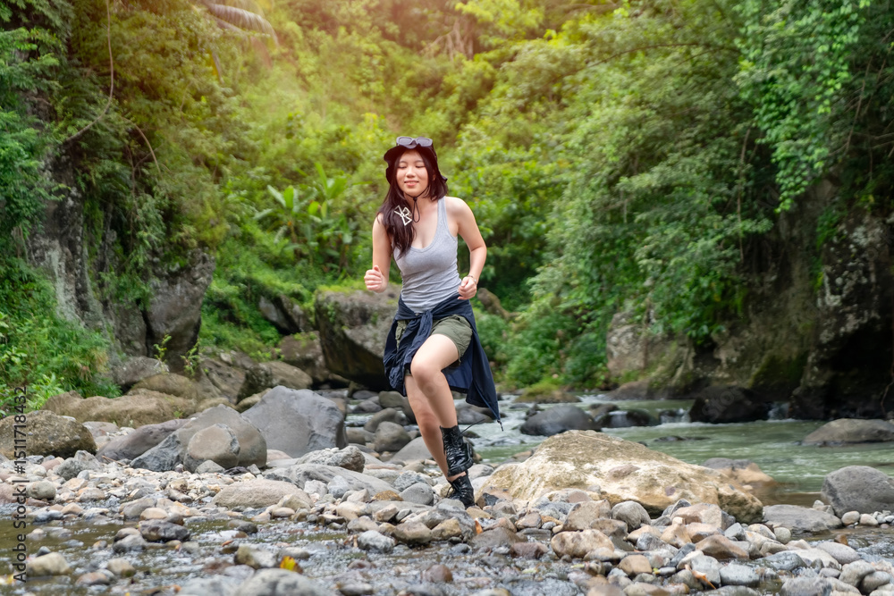Naklejka premium Asian Woman Running on Rocky Riverbed in Lush Green Jungle During Outdoor Adventure