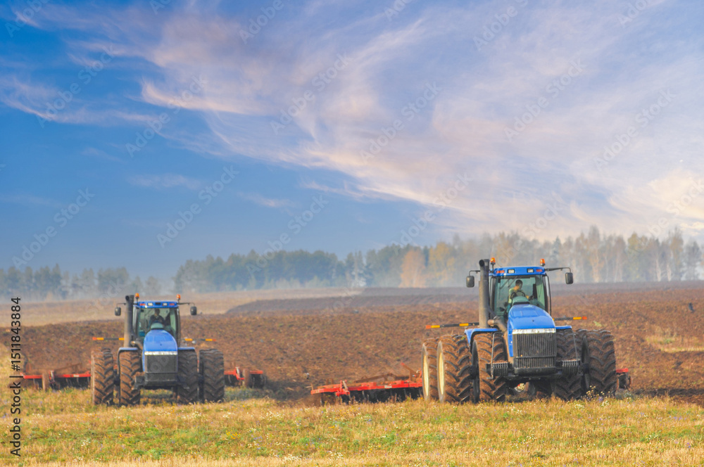 Fototapeta premium Two robust blue tractors cultivate a broad expanse of dark soil under swirling cloud formations, forging steady progress across open fields with autumn trees in the background.