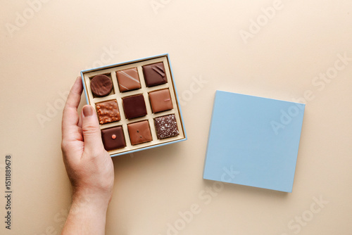Minimal flatlay of open box with chocolate squares and hand on beige background, elegant candy dessert packaging mockup concept
