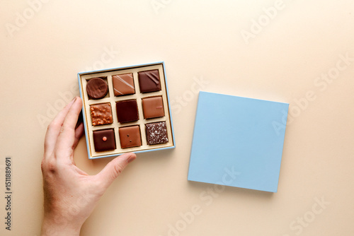 Minimal flatlay of open box with chocolate squares and hand on beige background, elegant candy dessert packaging mockup concept