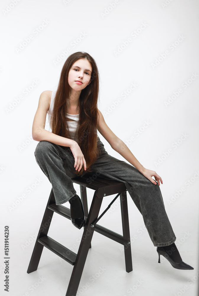 A girl in gray jeans and a white top is sitting on a chair on a white background. A girl in gray jeans and a white top is sitting on a chair on a white background.