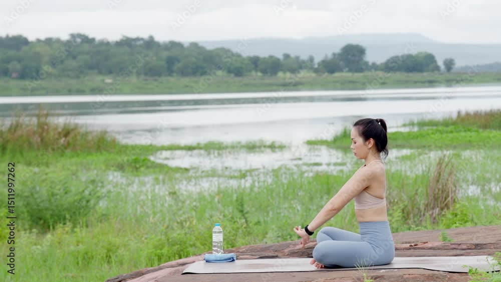 A woman is sitting on a mat by a lake. She is wearing a watch and a tank top. A bottle is next to her A woman is sitting on a mat by a lake. She is wearing a watch and a tank top. A bottle is next to her