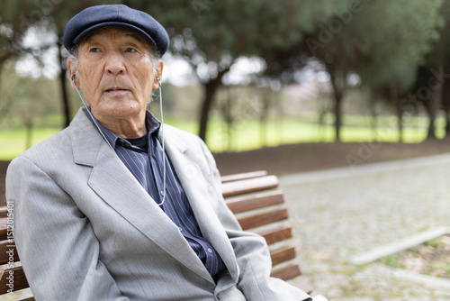 Elderly asian man listening music with earphones sitting on bench in park