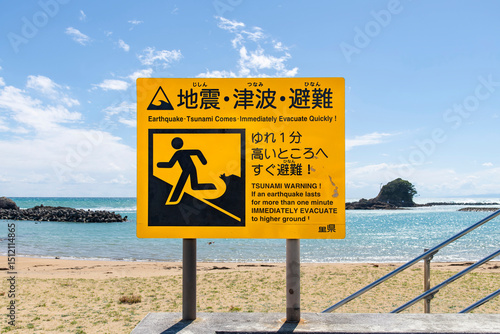 Large notice board on a beach in Japan warning for Tsunamis in case of an earthquake with sea, rock formation in shape of a wave and horizon in background (Japanese text translates on warning sign)