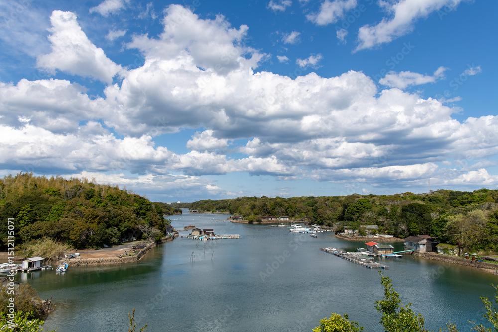 High level panoramic view over one of the bays of the Shima peninsula, Mie, Japan with small floating fishing settlements and large white cumulus clouds in blue sky High level panoramic view over one of the bays of the Shima peninsula, Mie, Japan with small floating fishing settlements and large white cumulus clouds in blue sky