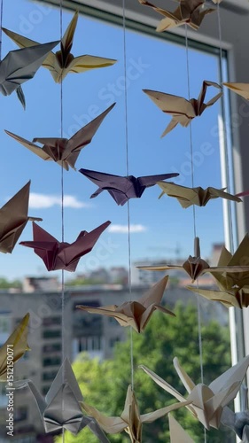 Hanging origami cranes of various colors by a window. Blue sky, trees, and city in the background. Sunlight highlights the figures' details.