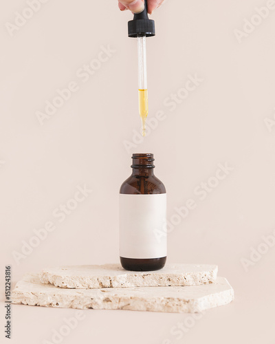 Glass amber dropper bottle on tan background with natural stones and blank label with dropper dripping oil