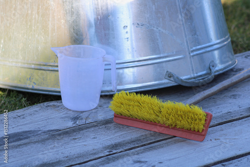 A plastic measuring cup and a yellow brush with a red handle placed on a wooden surface, with a metallic container in the background. The objects suggest practical use in a workshop or household setti