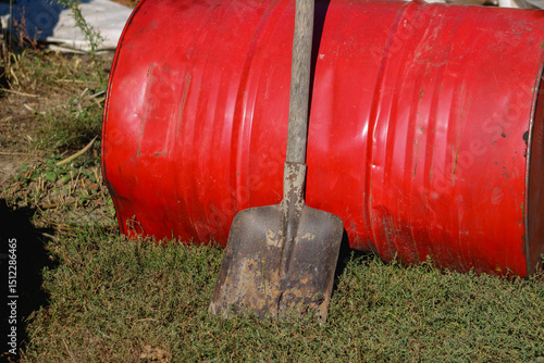 A red barrel with a shovel leaning against it, placed outdoors. The worn metal and utilitarian tools suggest practical, hands-on work	

