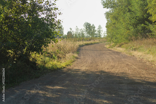 A winding dirt road surrounded by lush greenery, lined with trees and bushes. The natural pathway suggests a peaceful, outdoor scene