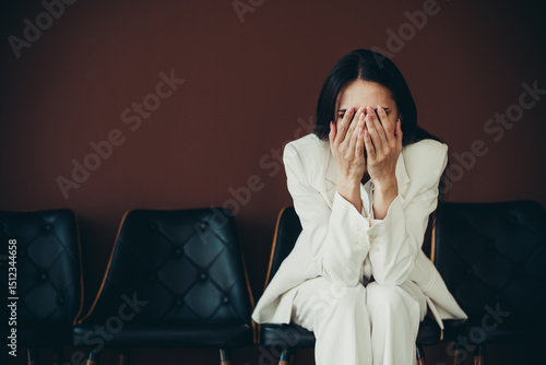 Slika na platnu Young professional woman in an elegant white suit sitting on a chair, covering her face with hands, in a modern workplace