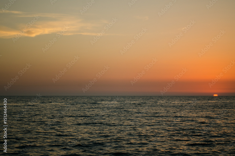 Fototapeta premium Calm ocean scene under warm, gradient sunset sky. Sun partially dips below horizon, casting orange glow. Peaceful atmosphere with gentle waves and expansive view