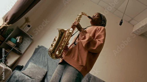 Young African American female musician playing solo jazz on saxophone while standing in front of smartphone on tripod and recording music video for social networks or online audience