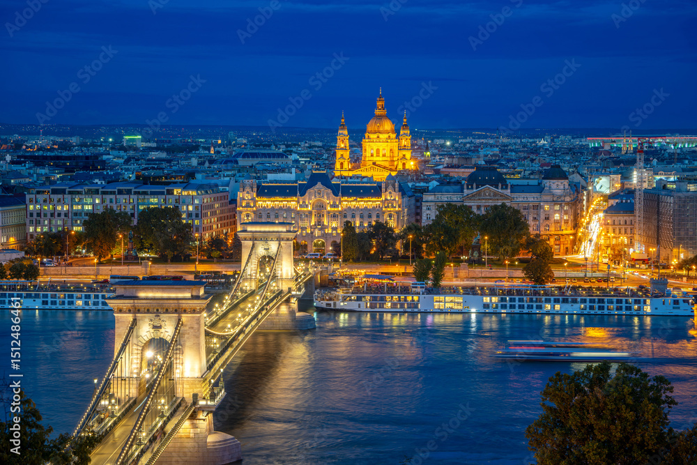 Obraz premium Night view of the Chain Bridge and St. Stephen's Basilica in Budapest, Hungary, illuminated by beautiful street lights, with boats gently drifting on the Danube River, showcasing the charm of the city