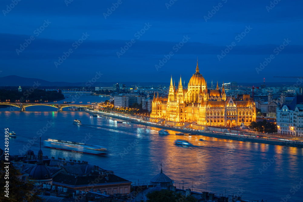 Obraz premium A beautiful night view of the Hungarian Parliament with boats on the Danube River, showcasing Budapest's vibrant lights and reflections