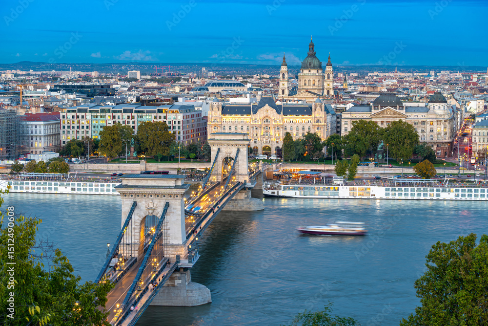 Obraz premium Beautiful view of the Chain Bridge and St. Stephen's Basilica in Budapest, Hungary, just before sunset, with street lights glowing and boats gently drifting on the Danube River