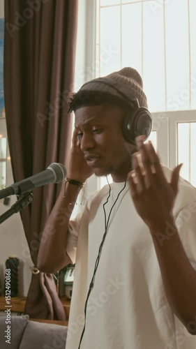 Vertical shot of young talented musician in casual attire and headphones standing in front of microphone in home studio and performing his new song during repetition