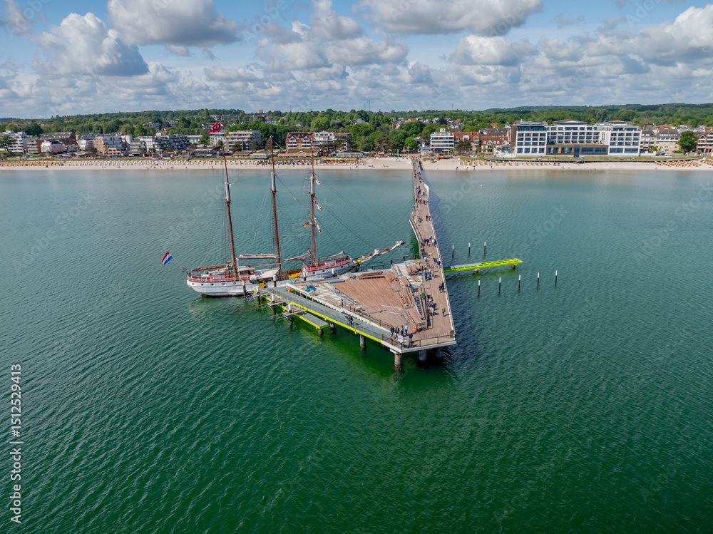 Obraz premium Aerial view of sailing ship docked at the long pier in Scharbeutz on the Baltic Sea. Wooden pier with three-masted sailing vessel at seaside resort.