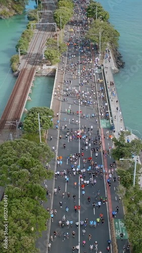 Aerial view of marathon runners on Tamaki Drive in Auckland, New Zealand. Spectators watch from a pedestrian bridge. The race promotes fitness and community engagement. PARNELL, AUCKLAND, NEW ZEALAND