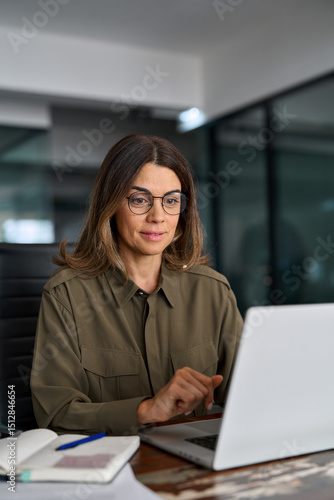 Busy mature middle aged professional business woman investor manager executive or lawyer attorney adviser looking at laptop at workplace working on computer in office managing business tasks.
