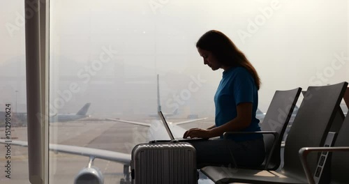 Young business woman sitting in waiting lounge chair at airport terminal, working on laptop to handle urgent tasks before her flight. Silhouetted against large window, passenger planes seen outdoors