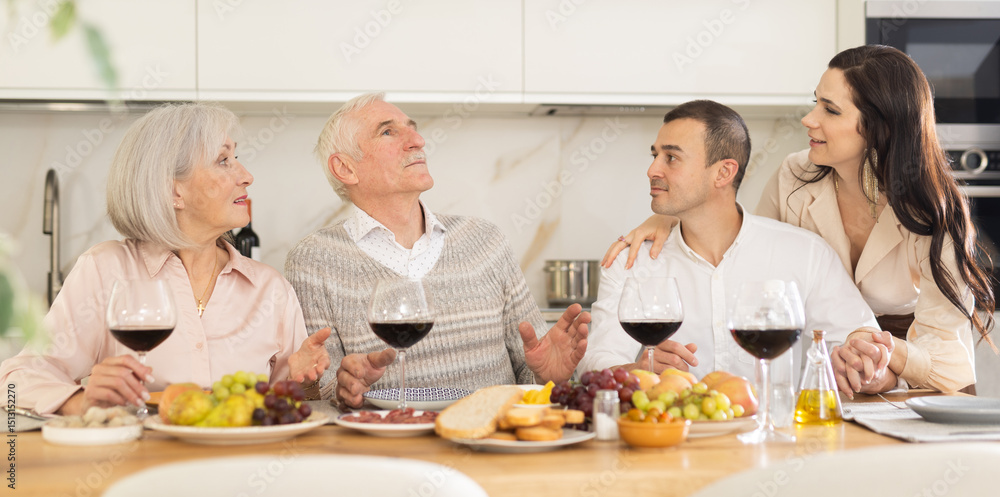 Man and his chosen one are sitting at table during family feast with elderly parents. Mature couple looks approvingly and gladly at their son and his girlfriend. Man and his chosen one are sitting at table during family feast with elderly parents. Mature couple looks approvingly and gladly at their son and his girlfriend.