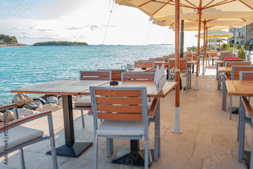 Bild auf Leinwand Tranquil seaside restaurant with modern wooden tables and chairs arranged under large umbrellas along a scenic waterfront promenade