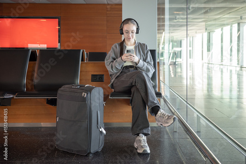 A young woman sits on a bench in a bright airport terminal, wearing headphones and looking at her smartphone with suitcase nearby. Waiting during international travel and digital technology.