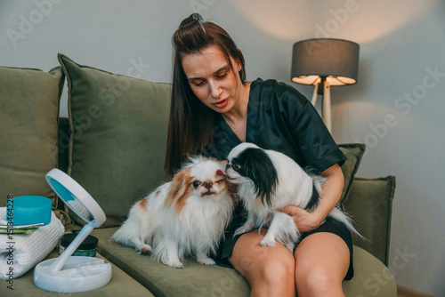 Canvas Print woman holds two japanese chin dogs as one licks other on green sofa in cozy home