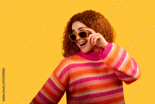 Cheerful redhead woman lowering sunglasses and smiling on yellow background