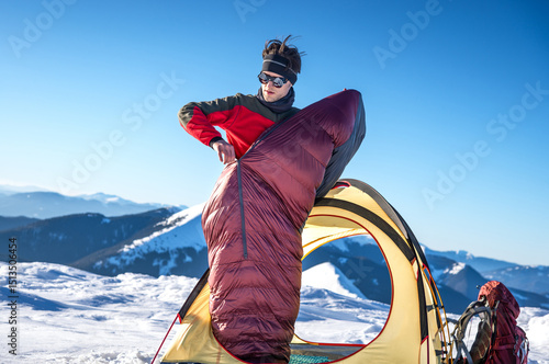 Tablou pe pânză Young man standing halfway out of a tent while zipping up a sleeping bag in a snowy mountain landscape