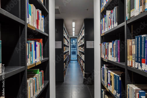 Photography A perspective shot down a long, narrow aisle in a modern library, flanked on both sides by tall bookshelves filled with various books