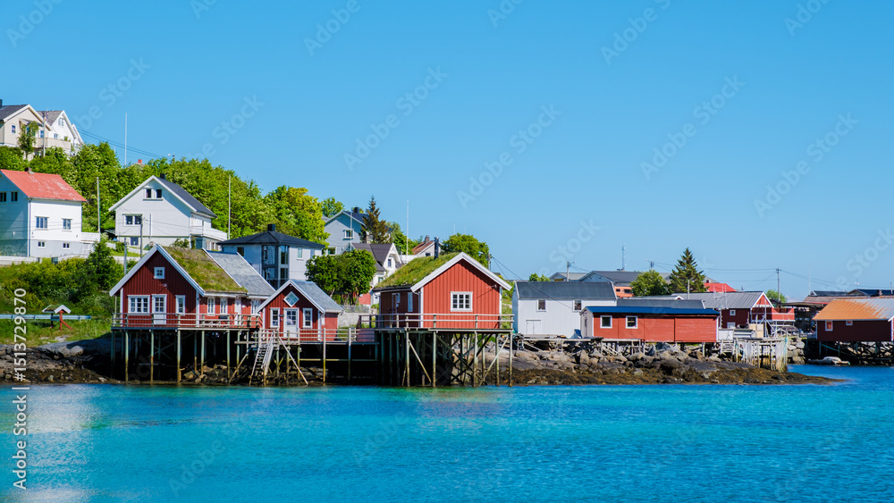 Naklejka premium Colorful wooden cabins line the tranquil waters of Lofoten Norway, embodying the scenic charm of coastal living. Surrounded by lush greenery, this picturesque spot invites relaxation and exploration.