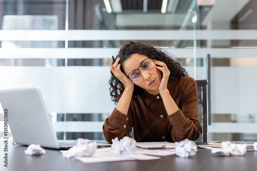 Obraz premium A frustrated businesswoman sits at her desk surrounded by crumpled paper, appearing overwhelmed. She looks tired after a long day.