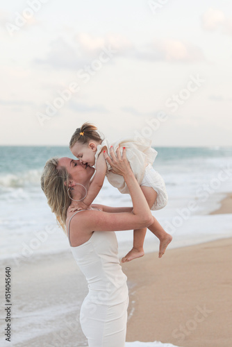 Beautiful young happy blonde girl in white short dress with her little daughter at sea on the beach in summer. Photo model. Model. Posing. Motherhood. Mom and daughter.