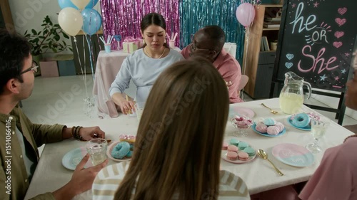 Medium arc shot of pregnant Caucasian woman enjoying dessert at gender reveal party, cheerful African American man stroking her belly, playfully teasing wife, and friends laughing