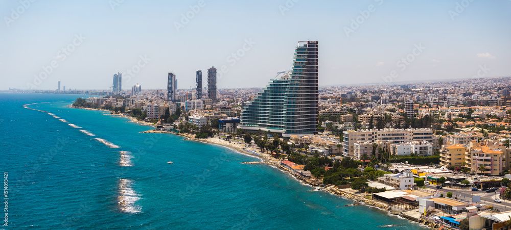 Fototapeta premium Aerial view of Limassol, Cyprus, featuring modern high rise buildings, a curved structure, turquoise sea, sandy beach, and wave breakers.