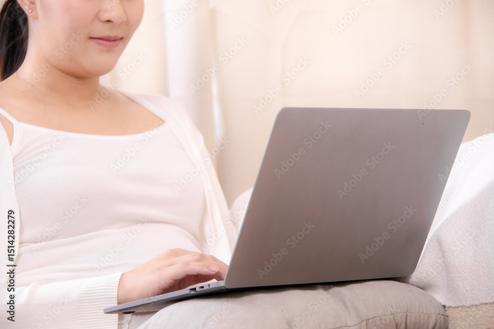 Fototapeta premium Pregnancy working, pregnant Asian woman using a laptop at home while gently touching her belly, combining productivity and care, small business owner, work-from-home, or maternity service promotions