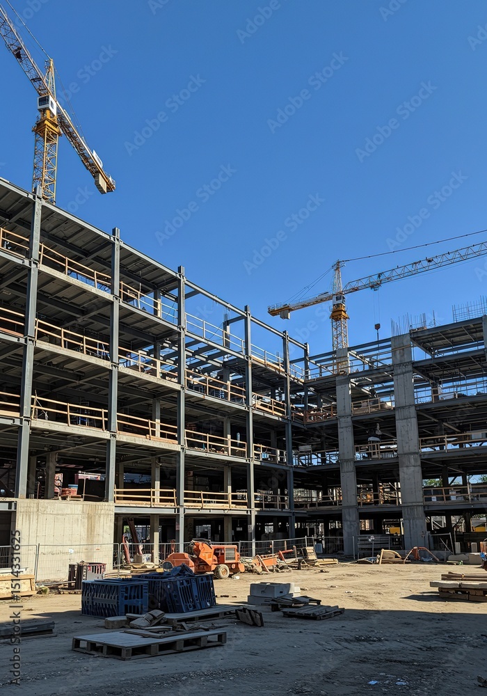 Naklejka premium Building under construction with steel beams and cranes against a clear blue sky.