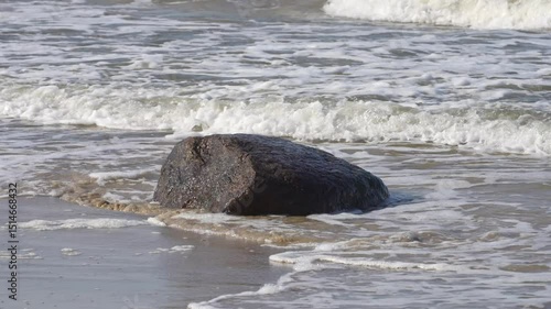 A large, dark coastal rock partially submerged by gentle sea waves on a sandy beach with white foam and sunlight reflections