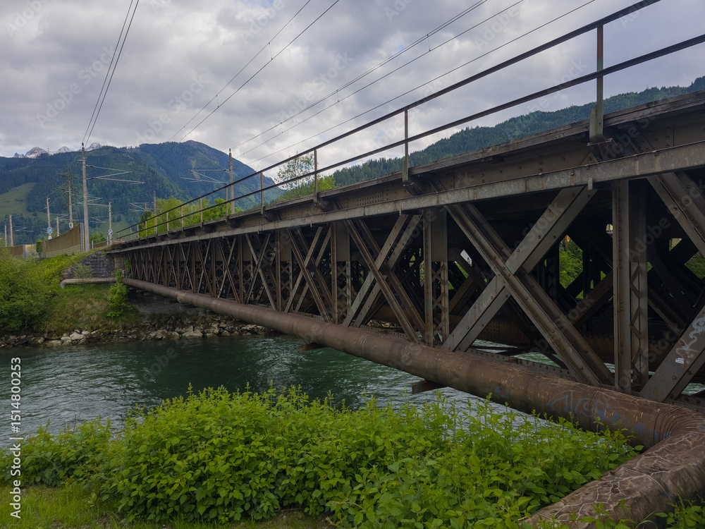 Fototapeta premium A sturdy metal bridge spans a flowing river, with scenic mountains in the distance, under a cloudy sky