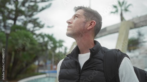 Wallpaper Mural Young man standing on a city street, wearing a black vest, looking thoughtful, surrounded by urban greenery and palm trees, capturing a modern and casual outdoor vibe. Torontodigital.ca