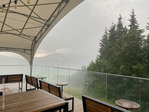 A mountaintop cafe with wooden tables and benches during a heavy rain in the mountains. Rain drops dripping down the glass fence. Silhouettes of mountains and forest in the fog in the background.